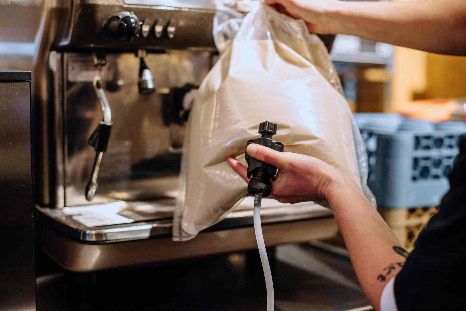 Une personne relie un grand sac en plastique rempli d'un liquide crémeux à une machine à expresso commerciale à l'aide d'un tuyau et d'une buse.