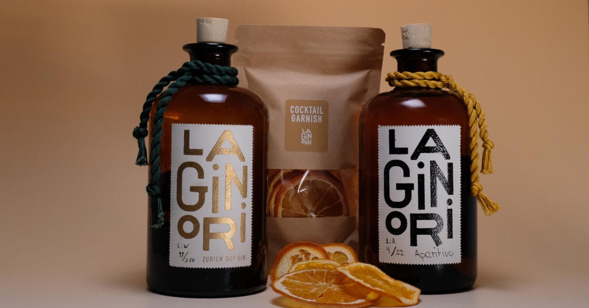 Zwei braune Glasflaschen mit der Aufschrift „LAGINORI“ und dekorativen Kordeln stehen neben einem braunen Beutel mit der Aufschrift „Cocktail Garnish“, gefüllt mit getrockneten Orangenscheiben, auf neutralem Hintergrund. Die getrockneten Orangenscheiben sind im Vordergrund platziert.