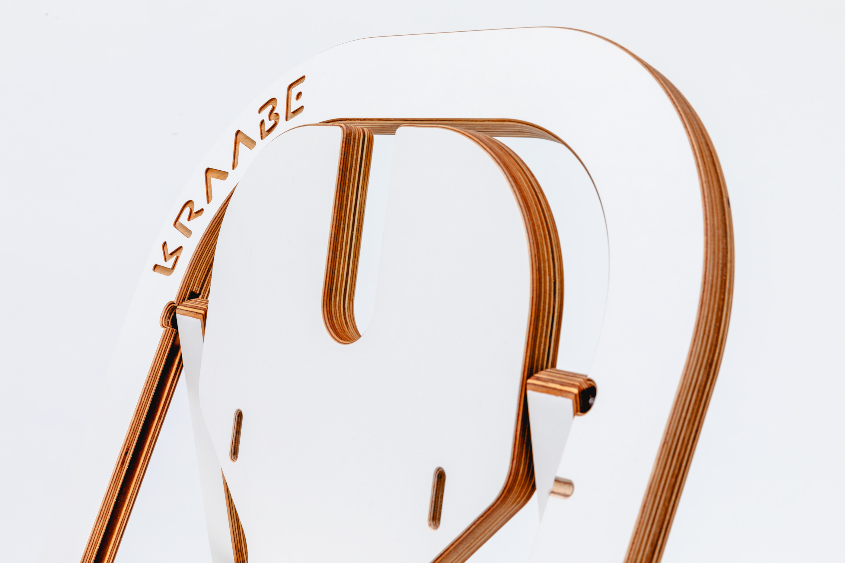 Gros plan sur une chaise en bois moderne et minimaliste, avec des surfaces blanches et des bords en bois naturel. Le nom de la marque KRAABE est gravé sur le bord supérieur du dossier de la chaise. L'arrière-plan est blanc.