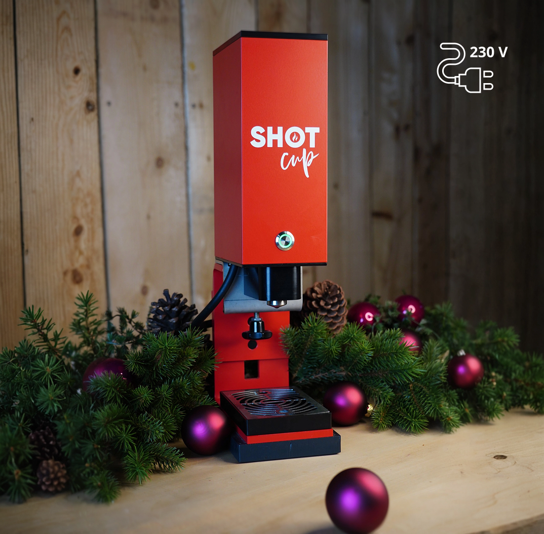 Eine rote Espressomaschine mit der Aufschrift SHOT cup steht auf einem Holztisch, der mit Tannenzweigen und lila Ornamenten verziert ist. In der rechten oberen Ecke ist ein 230-V-Stromsymbol abgebildet. Holzbretter bilden den Hintergrund.