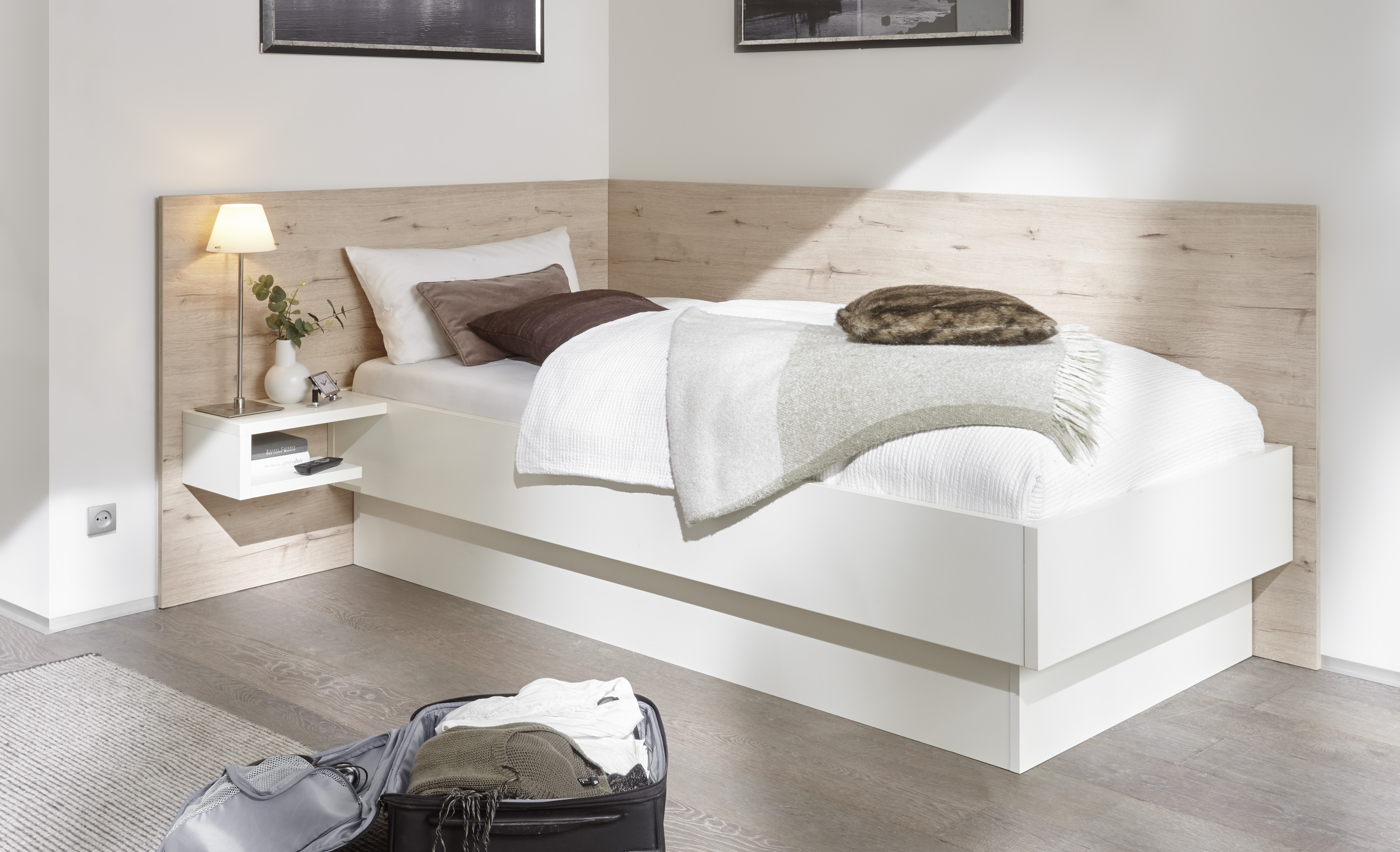 Ein ordentlich gemachtes Einzelbett mit weißem und beigem Bettzeug steht in einer Ecke mit einem hölzernen Kopfteil. Auf einem kleinen Regal stehen eine Lampe und Dekoration. Ein offener Koffer steht auf dem Holzboden neben einem Teppich.