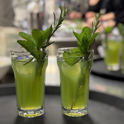 Zwei Gläser, gefüllt mit einem grünen Getränk, garniert mit frischen Minze- und Rosmarinzweigen, stehen auf einem dunklen Tablett. Der Hintergrund ist sanft verschwommen, mit undeutlichen Figuren und hellen Farben.