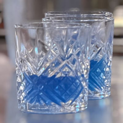 Zwei klare, gemusterte Glasbecher, die teilweise mit einer blauen Flüssigkeit gefüllt sind, stehen dicht nebeneinander auf einer spiegelnden Fläche mit unscharfem Hintergrund.