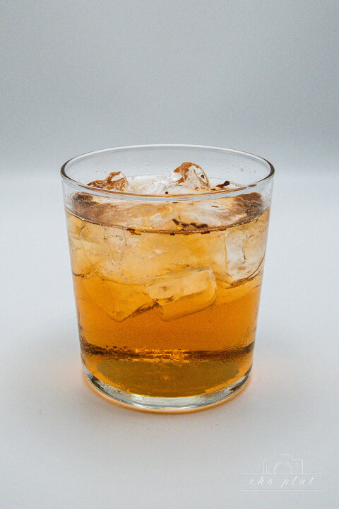 Un verre clair rempli d'un liquide ambré et de glaçons, sur un fond blanc sobre.