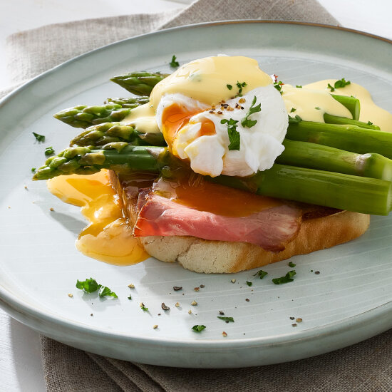 Une assiette de pain grillé, recouverte de jambon, de bâtonnets d'asperges, d'un œuf poché au jaune d'œuf liquide et d'une sauce hollandaise crémeuse, le tout garni de persil haché et de poivre noir.