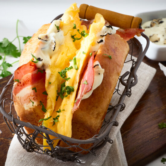 Ein Sandwich mit fluffigem Rührei, Schinken und einer cremigen Sauce, garniert mit gehackten Kräutern, serviert in einem rustikalen Drahtkorb auf einer Stoffserviette. Im Hintergrund stehen eine kleine Schale mit Aufstrich und frischer Petersilie.