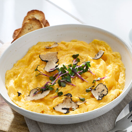 Eine Schale mit cremigem Rührei, garniert mit dünnen Trüffelscheiben und Mikrogrün, serviert mit getoasteten Brotscheiben als Beilage.