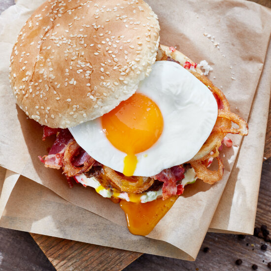 Un petit pain hamburger au sésame, garni d'un œuf au plat, de rondelles d'oignons croustillantes, de bacon et de sauce, servi sur du papier parchemin brun.
