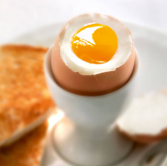 Un œuf à la coque avec un jaune liquide est assis dans un coquetier, avec en arrière-plan des tranches de pain grillées sur une assiette blanche.