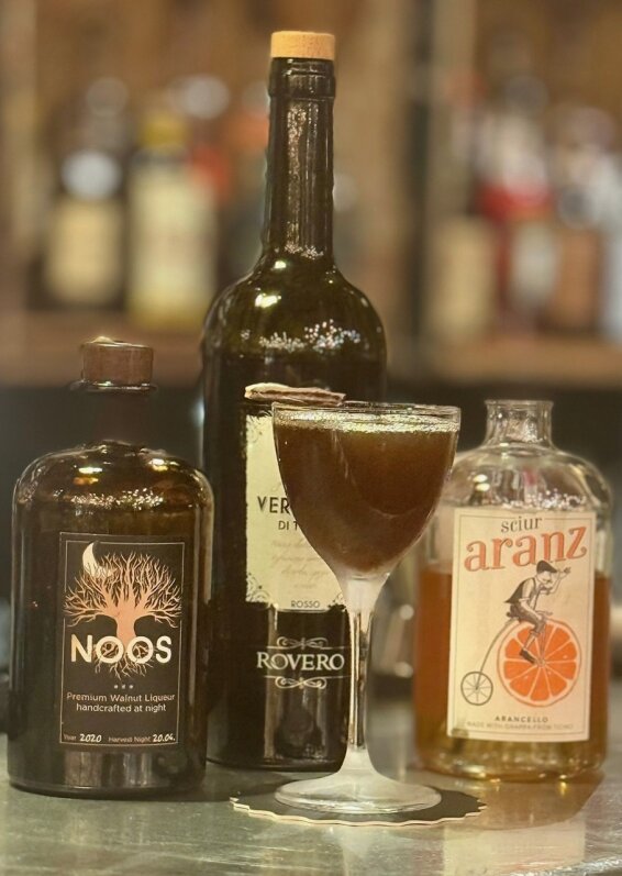 Un cocktail sombre dans un verre coupe est posé sur un bar, entouré de bouteilles de liqueur de noix NOOS, de vermouth Rosso Rovero et d'eau-de-vie d'orange Scür Aranz. L'arrière-plan est doucement flou.