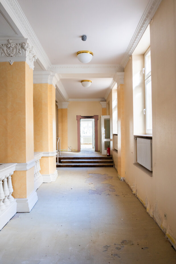 Ein heller, leerer Flur mit gelb gestrichenen Wänden, weißer Verkleidung und zwei Deckenleuchten. Durch die Fenster auf der rechten Seite strömt Sonnenlicht. Der Boden scheint abgenutzt zu sein, und es gibt Stufen, die zu einer Tür am anderen Ende führen.