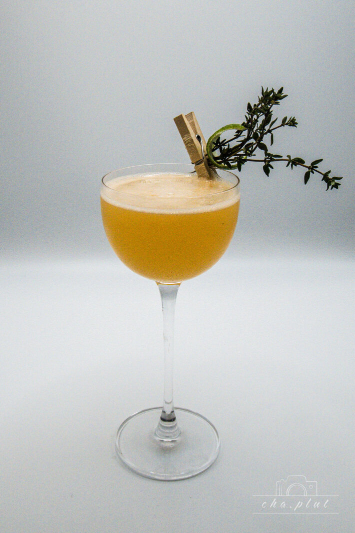 Un cocktail jaune dans un verre coupe, garni d'une branche de thym et d'une branche de citron vert maintenue par une petite pince à linge en bois, sur un fond clair et sobre.