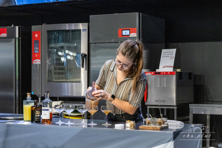 Une femme portant des lunettes et une chemise à rayures verse délicatement une boisson dans un verre à cocktail à un comptoir de bar devant lequel sont disposés des bouteilles, des mixeurs et des verres à cocktail. En arrière-plan, on peut voir des ustensiles de cuisine en acier inoxydable.