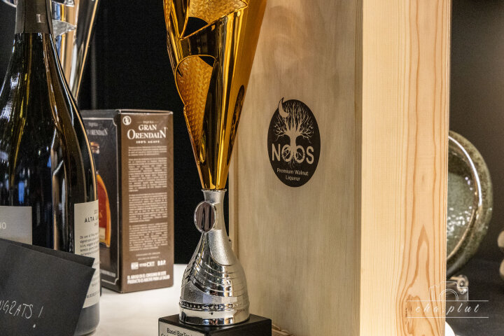 Un trophée doré et argenté est posé sur une étagère à côté de bouteilles de vin et d'une caisse en bois portant l'inscription NOOS Premium Liqueur de noix, tandis qu'en arrière-plan, on peut voir différentes boissons emballées.