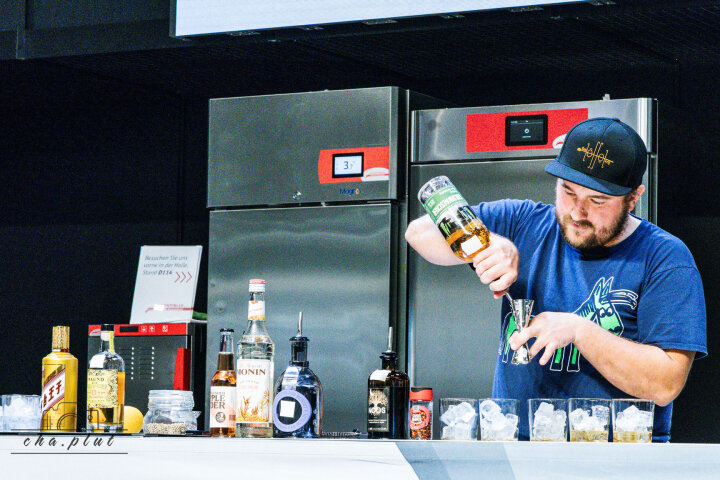 Derrière un comptoir de bar, un barman en chemise et casquette bleues verse une boisson verte dans un jigger. Devant lui se trouvent plusieurs bouteilles et verres remplis de glace et, en arrière-plan, des réfrigérateurs en acier inoxydable.
