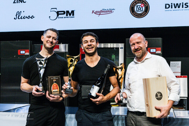 Lors d'une remise de prix, trois hommes se tiennent debout, souriants, tenant des trophées et des bouteilles à la main, tandis que des logos de sponsors s'affichent derrière eux sur l'écran. Un homme tient une caisse en bois, un autre un trophée en verre et une bouteille.