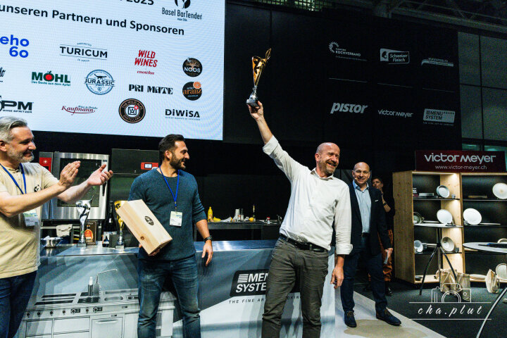 Un homme souriant, vêtu d'une chemise blanche, soulève solennellement un trophée, tandis que trois autres hommes se tiennent à proximité. Derrière eux, on peut voir un grand écran avec les logos des sponsors et des partenaires.