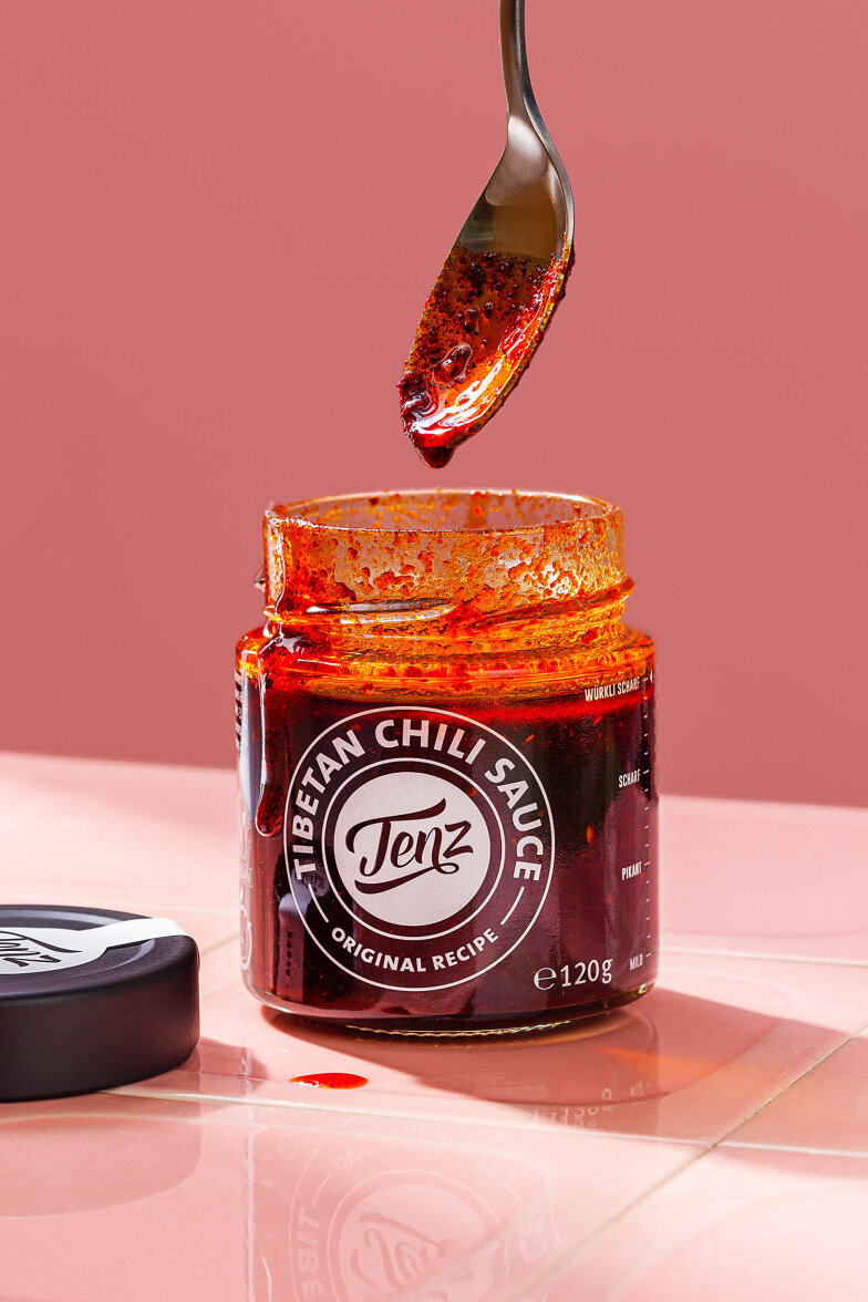 Ein Glas Tenz Tibetan Chili Sauce steht ohne Deckel auf einer rosa Fläche. Ein Löffel mit der roten, öligen Sauce schwebt über dem offenen Glas, und etwas Sauce ist auf die Oberfläche darunter getropft.