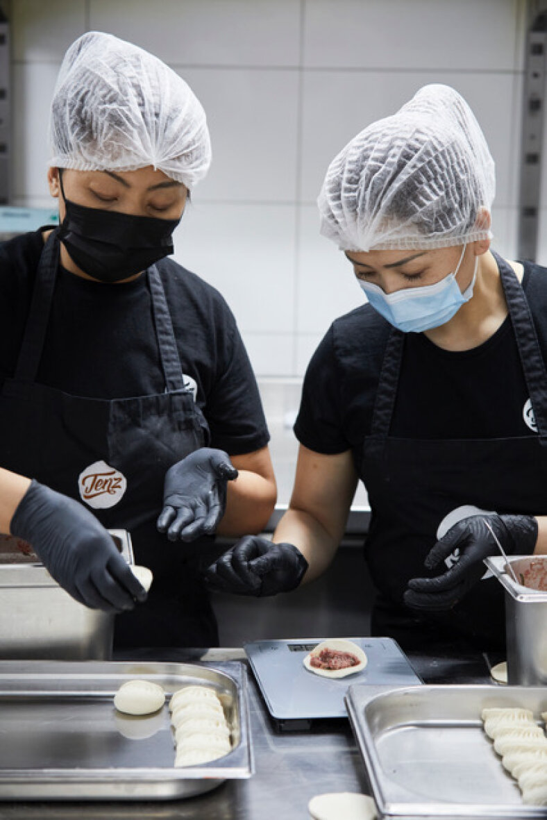 Deux personnes portant des résilles, des masques faciaux et des gants composent des boulettes dans une cuisine, en ajoutant la farce sur la pâte et en la pesant sur une balance numérique. Des plateaux de boulettes prêtes à l'emploi sont disposés sur le comptoir.