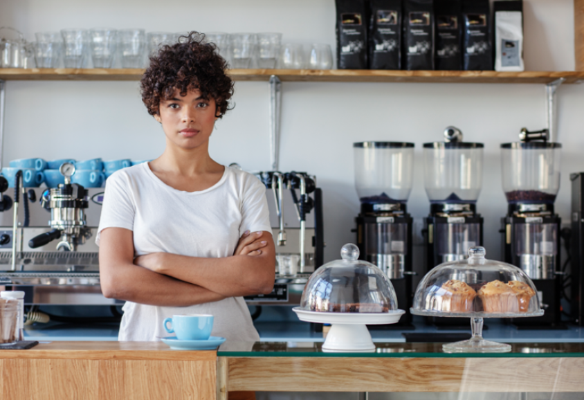 Une personne portant un t-shirt blanc se tient les bras croisés derrière le comptoir d'un café, avec en arrière-plan une machine à café, des moulins à café et des produits de boulangerie présentés sous des coupoles en verre.