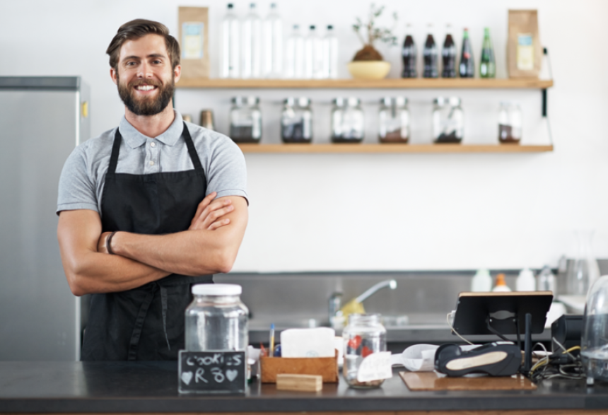 Un homme souriant, barbu et portant un tablier, se tient derrière le comptoir d'un café ou d'une cafétéria moderne, avec en arrière-plan des bocaux, une caisse enregistreuse, des étagères de bouteilles et des plantes.