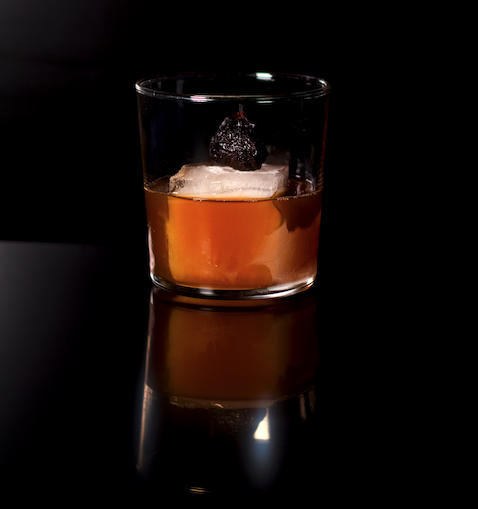 Un verre à cocktail ambré avec un gros glaçon clair et une garniture de cerises foncées sur une surface noire réfléchissante.