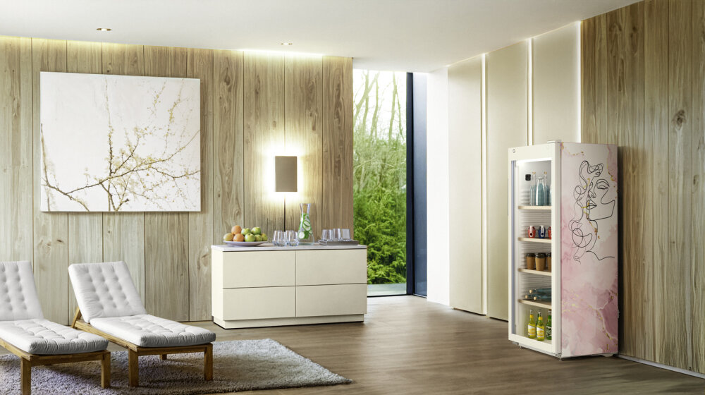 Ein modernes Wohnzimmer mit holzgetäfelten Wänden, einer weißen Anrichte, zwei Loungesesseln, einem großen abstrakten Gemälde und einem offenen Kühlschrank mit rosafarbenem Blumenmuster, in dem sich Getränke und Speisen befinden. Eine Glastür gibt den Blick auf die grüne Umgebung frei.