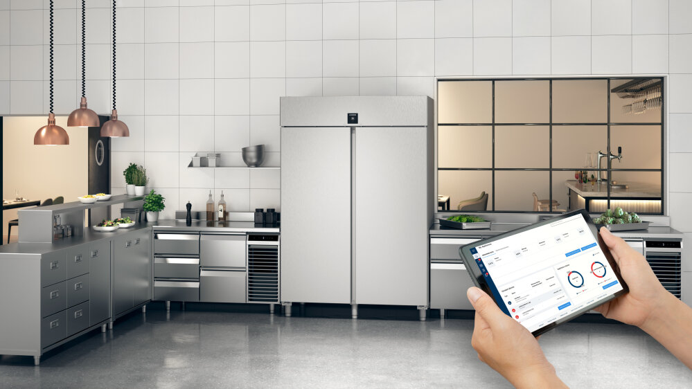 Une cuisine industrielle moderne avec des appareils et des comptoirs en acier inoxydable ; au premier plan, des mains tiennent une tablette sur laquelle sont affichés des diagrammes et des données.