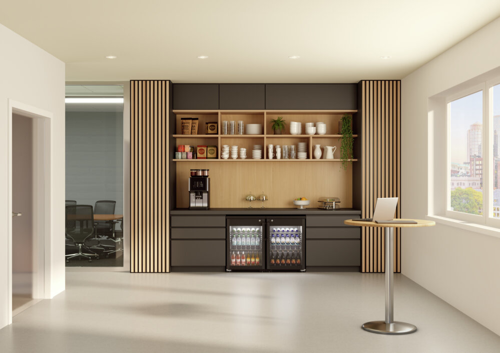 Moderne Büro-Küchenzeile mit dunklen Schränken, offenen Holzregalen mit Tassen und Tellern, einer Kaffeemaschine, zwei Kühlschränken mit Glastüren, in denen sich Getränke befinden, und einem kleinen runden Tisch mit einem Laptop neben einem großen Fenster.
