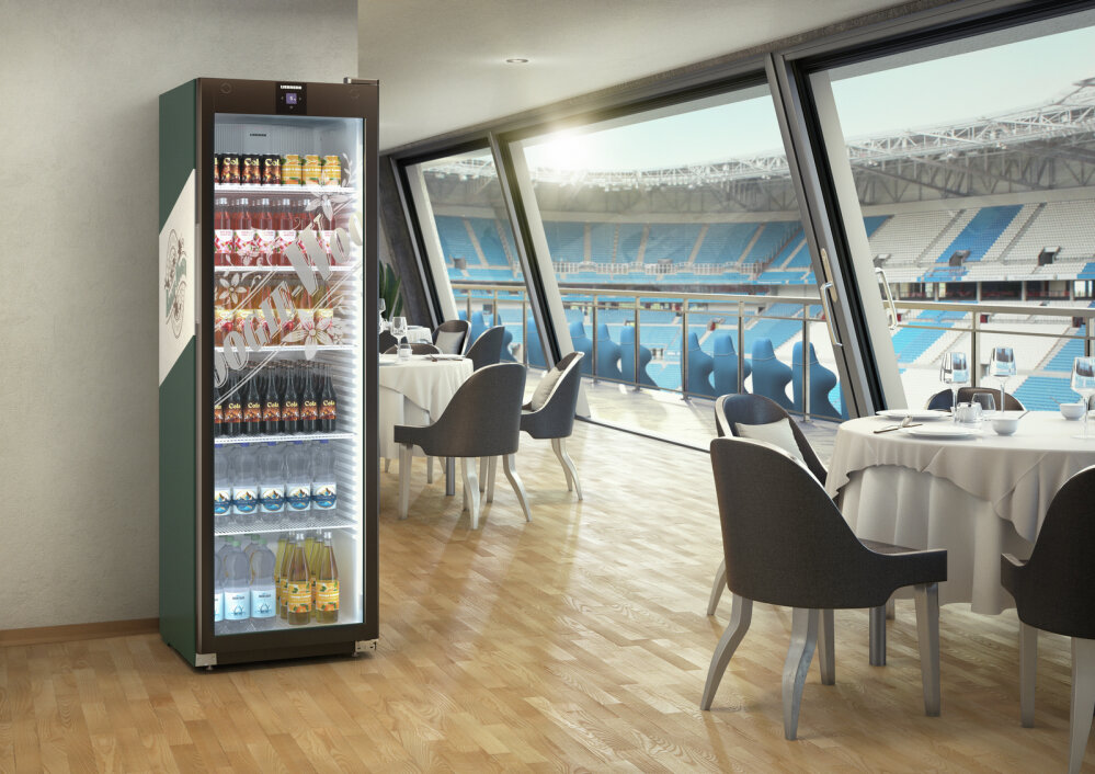 Ein Kühlschrank mit Glastüren, gefüllt mit Getränken und Snacks, steht in einem modernen Essbereich mit runden Tischen und Stühlen, neben großen Fenstern mit Blick auf ein Stadion mit blauen Sitzen.