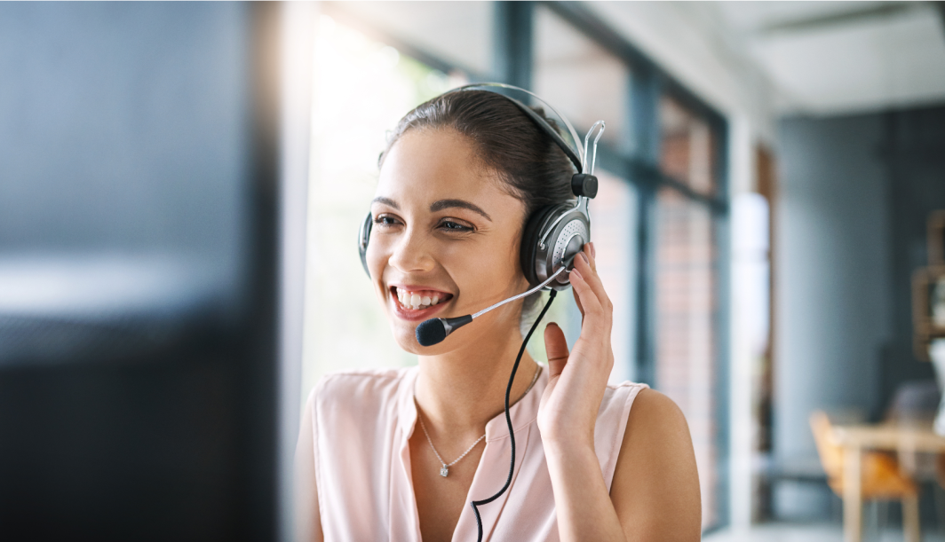 Une femme souriante portant un casque est assise à un bureau dans un bureau lumineux et regarde un écran d'ordinateur lorsqu'elle parle, ce qui suggère qu'elle travaille dans le service clientèle ou dans un centre d'appels.