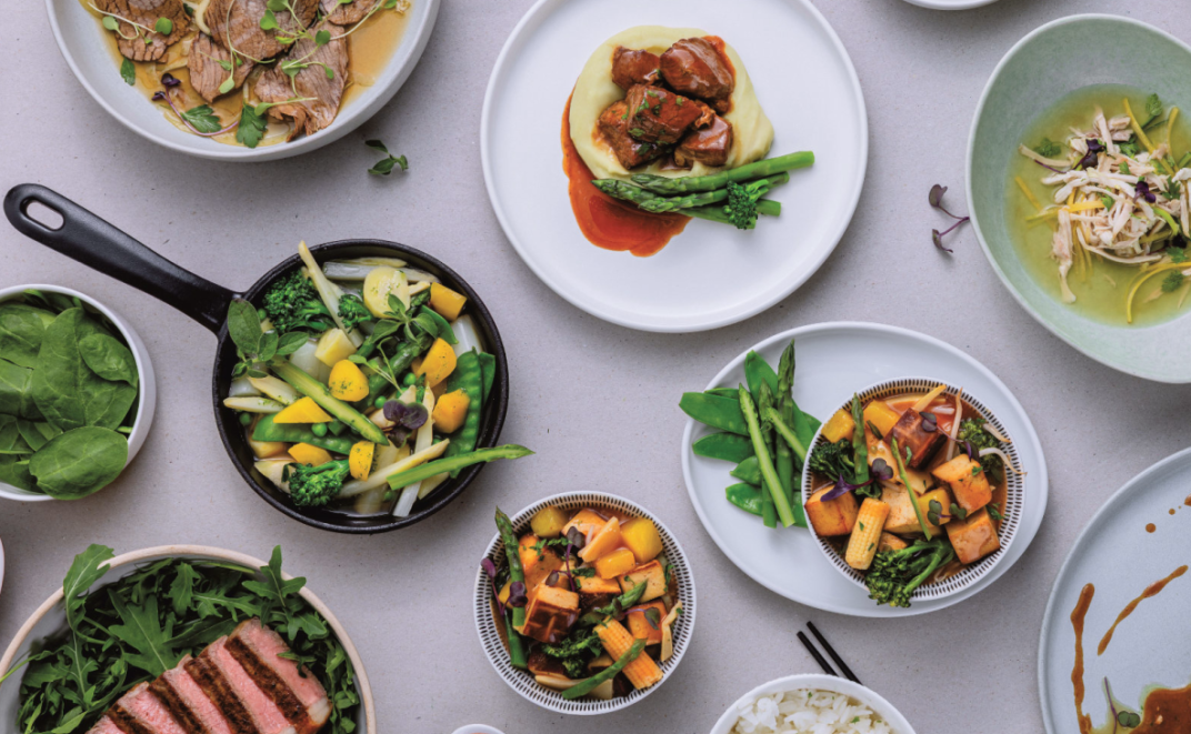 Eine bunte Auswahl an Gerichten, darunter gegrilltes Fleisch, gebratenes Gemüse, Kartoffelpüree mit Spargel und Soße, frisches Grün und Suppe, auf einem hellen Tisch angerichtet.