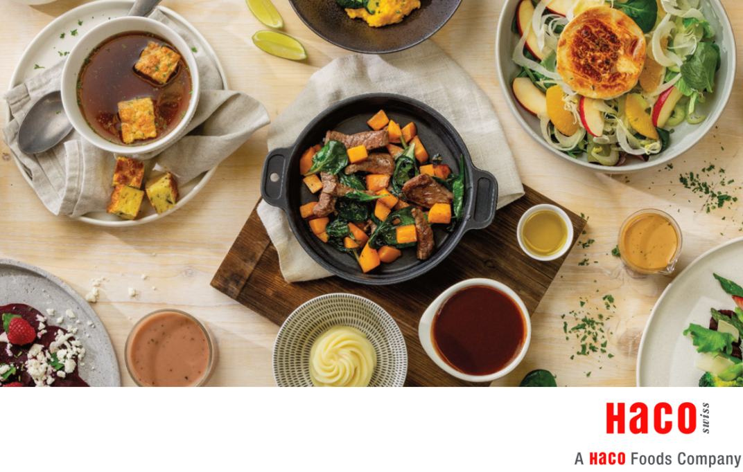 Vue aérienne d'une table avec divers plats, dont une poêle de bœuf avec des légumes, de la soupe, des salades, des sauces et de la purée de pommes de terre. Le logo de la société HACO Foods apparaît en bas de l'image.