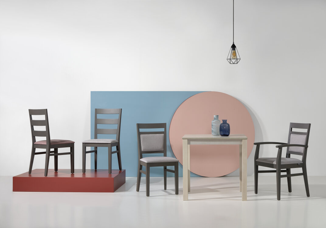 Vier Holzstühle, ein kleiner quadratischer Tisch mit zwei Vasen und eine hängende Glühbirne sind in einem modernen Studio mit einem roten Podest und einem blauen und rosa geometrischen Hintergrund angeordnet.