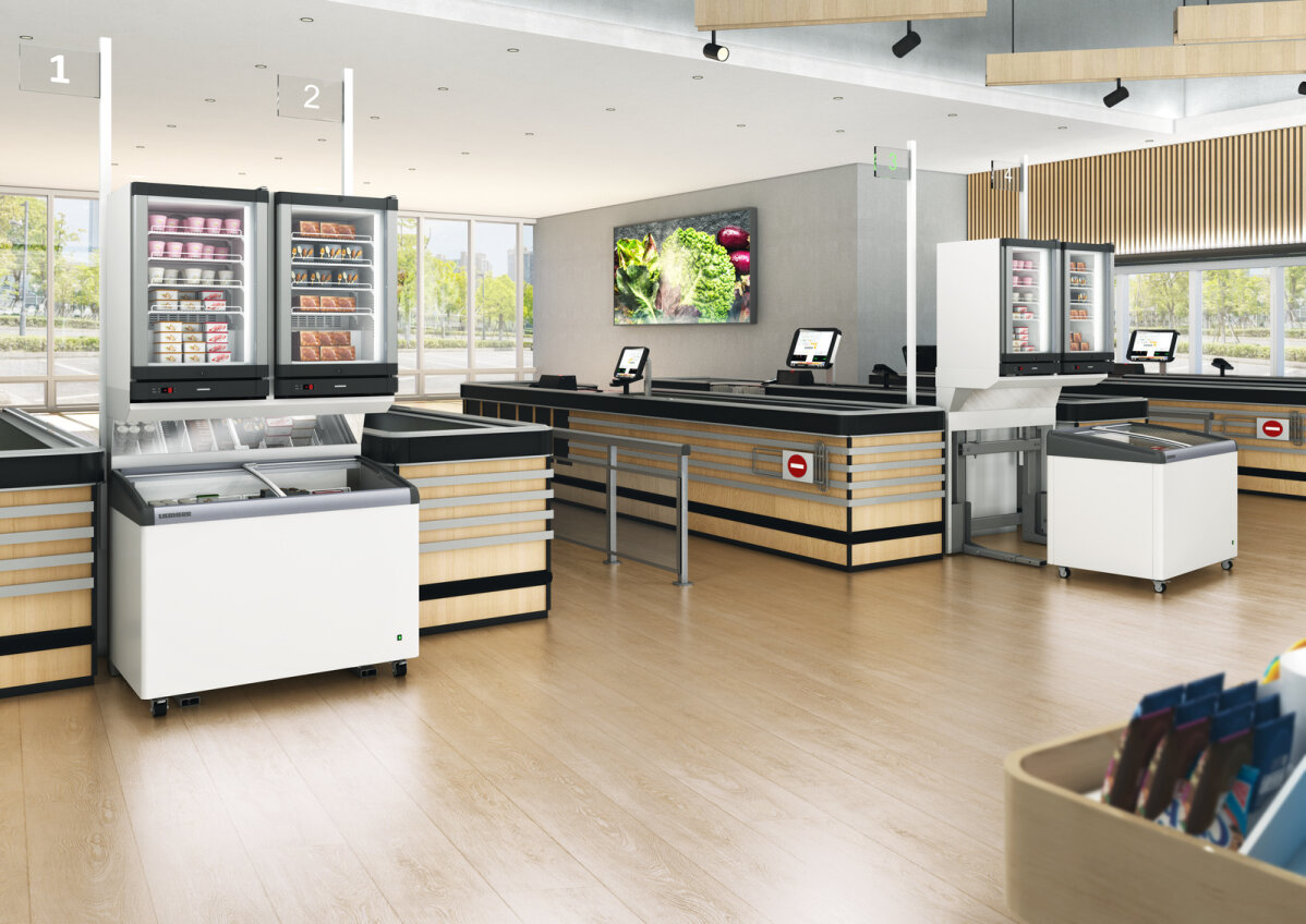 Moderner Kassenbereich eines Lebensmittelgeschäfts mit Holzakzenten, Self-Checkout-Stationen, mit Lebensmitteln gefüllten Tiefkühltruhen und digitalen Bildschirmen; an der Wand eine Gemüseauslage und im Vordergrund Snackregale.