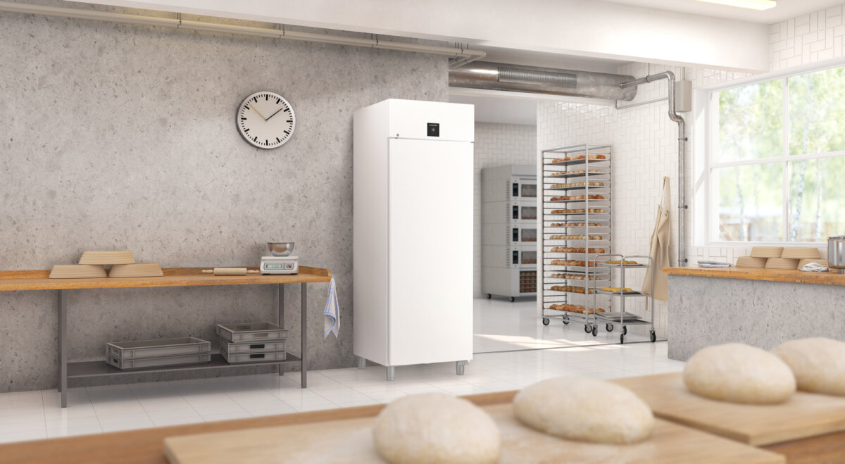 Eine helle Bäckereiküche mit Teigkugeln auf einer Holztheke im Vordergrund, einer Uhr an einer grauen Wand, einem hohen weißen Kühlschrank, Backständern mit Brottabletts und Sonnenlicht, das durch große Fenster fällt.