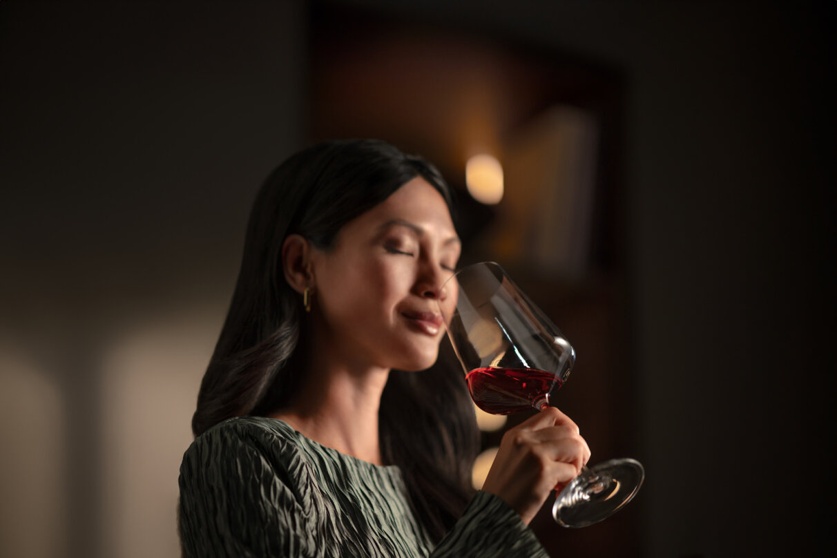 Une femme aux longs cheveux bruns tient un verre de vin rouge près de son visage, les yeux fermés, et semble en apprécier l'arôme, dans un intérieur faiblement éclairé.
