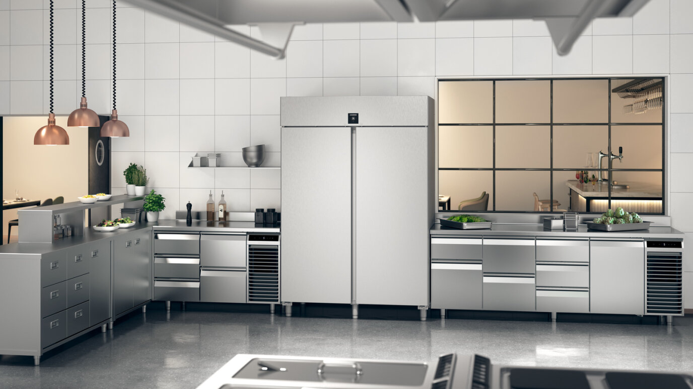 Eine moderne Großküche mit Geräten, Arbeitsflächen und Schränken aus Edelstahl, mit einem großen Kühlschrank in der Mitte und verschiedenen Küchengeräten und frischen Zutaten, die übersichtlich angeordnet sind.