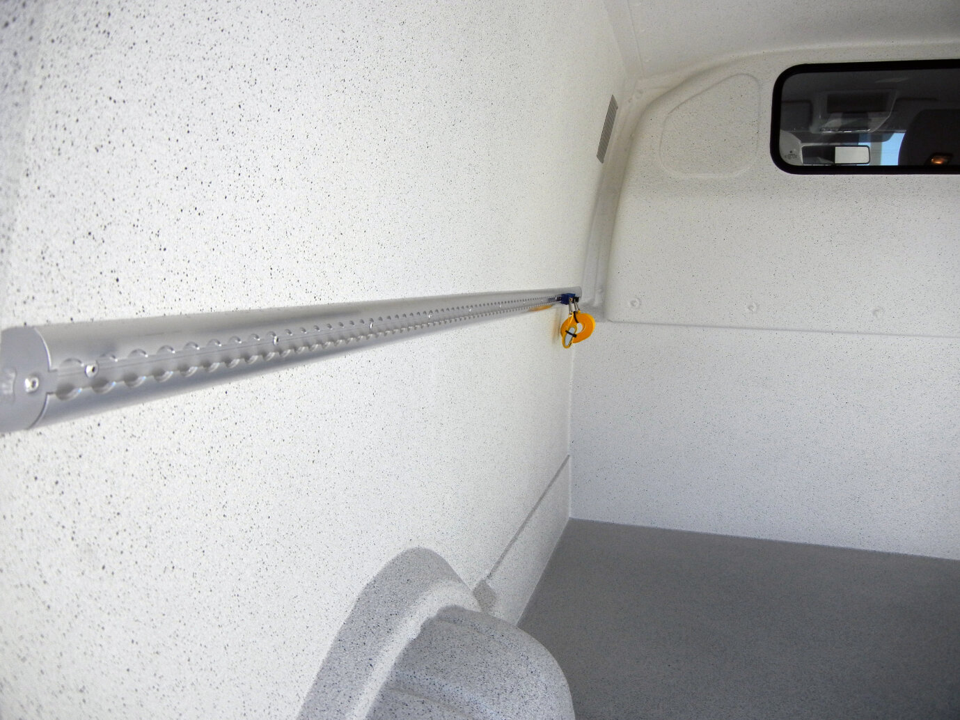 L'intérieur d'un camion blanc et vide, avec un mur moucheté, un sol gris, un rail métallique horizontal sur le côté gauche et une petite fenêtre sur le côté droit, près du plafond.