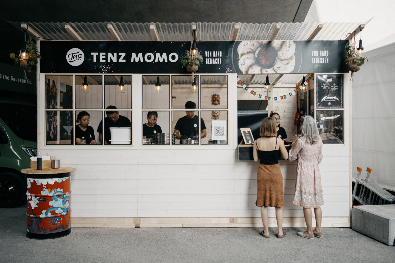 Ein weißer Imbissstand namens TENZ MOMO mit fünf Angestellten im Inneren bedient zwei Kundinnen. Bunte Fahnen schmücken die Fassade, und links steht eine dekorative Trommel. Die Atmosphäre wirkt locker und einladend.