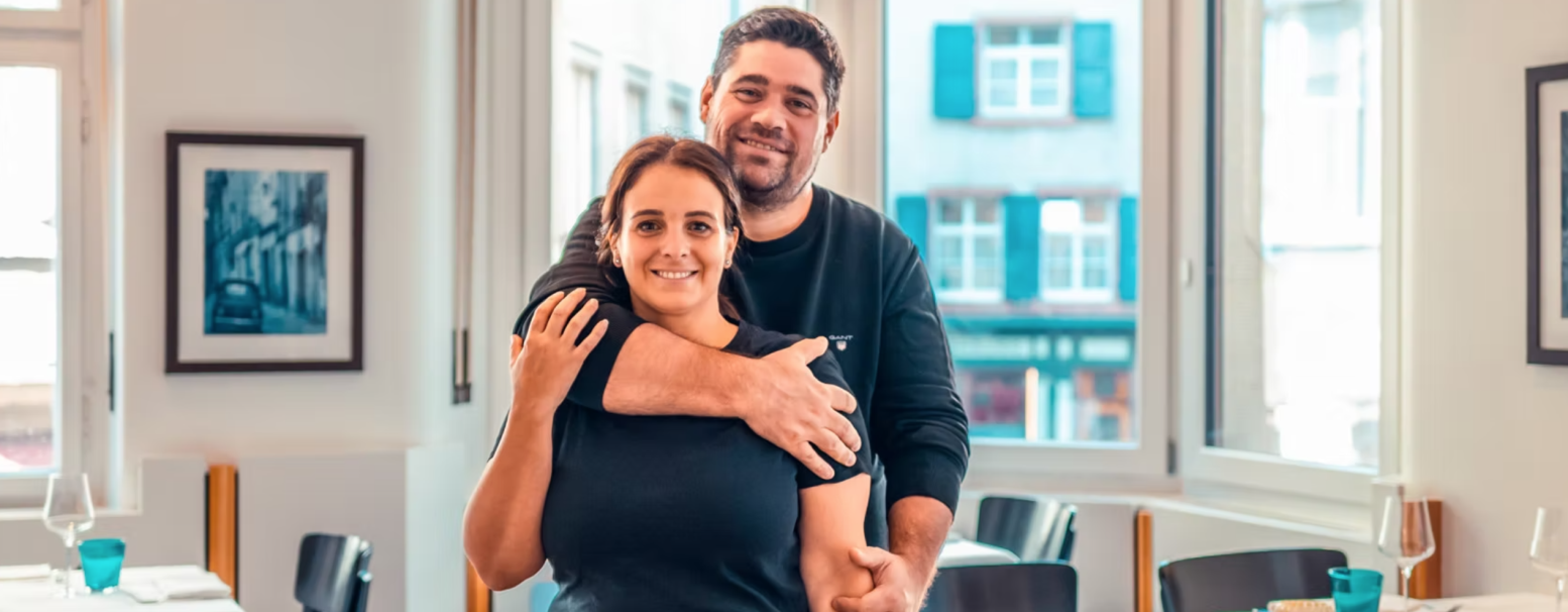 Un homme se tient derrière une femme, souriant, les bras autour de ses épaules, à l'intérieur d'un restaurant lumineux avec de grandes fenêtres, des photos encadrées au mur et des tables dressées à l'arrière-plan.