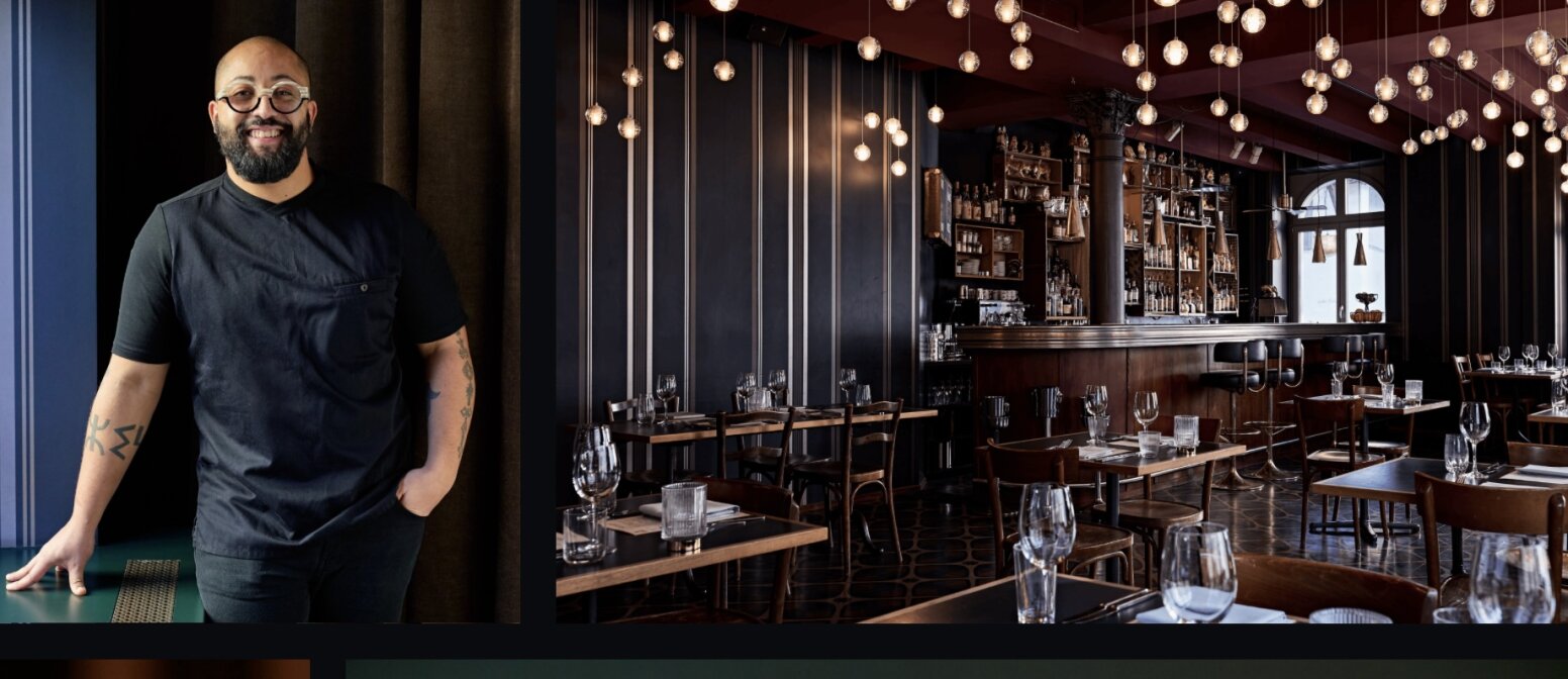 Un homme souriant, portant des lunettes et des tatouages, se tient devant un rideau sombre. À côté de lui se trouve un restaurant moderne avec des tables en bois équipées de verres, des murs sombres, des lampes suspendues et un bar bien approvisionné.