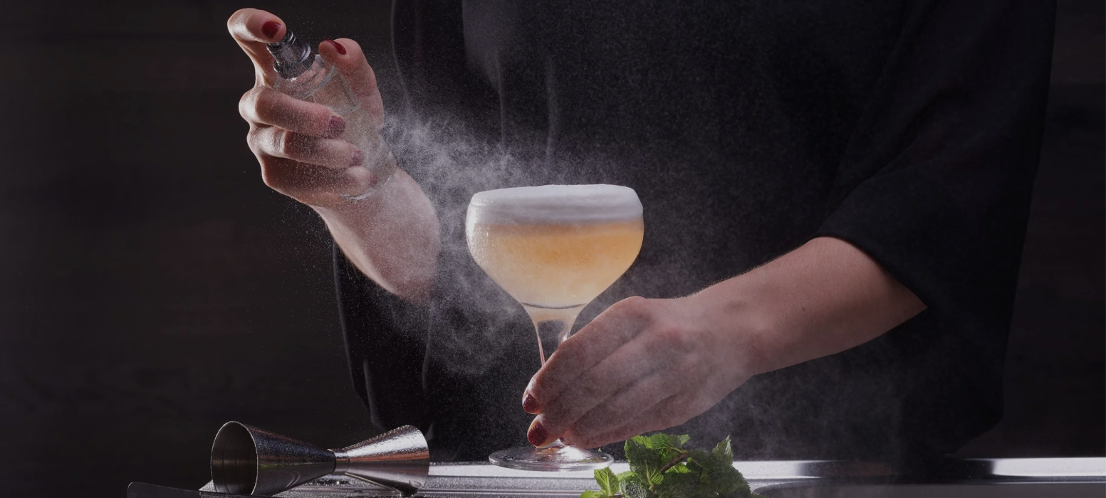Une personne vaporise une fine brume sur un cocktail mousseux dans un verre coupe, avec un jigger et des feuilles de menthe fraîches sur la table, sur un fond sombre.