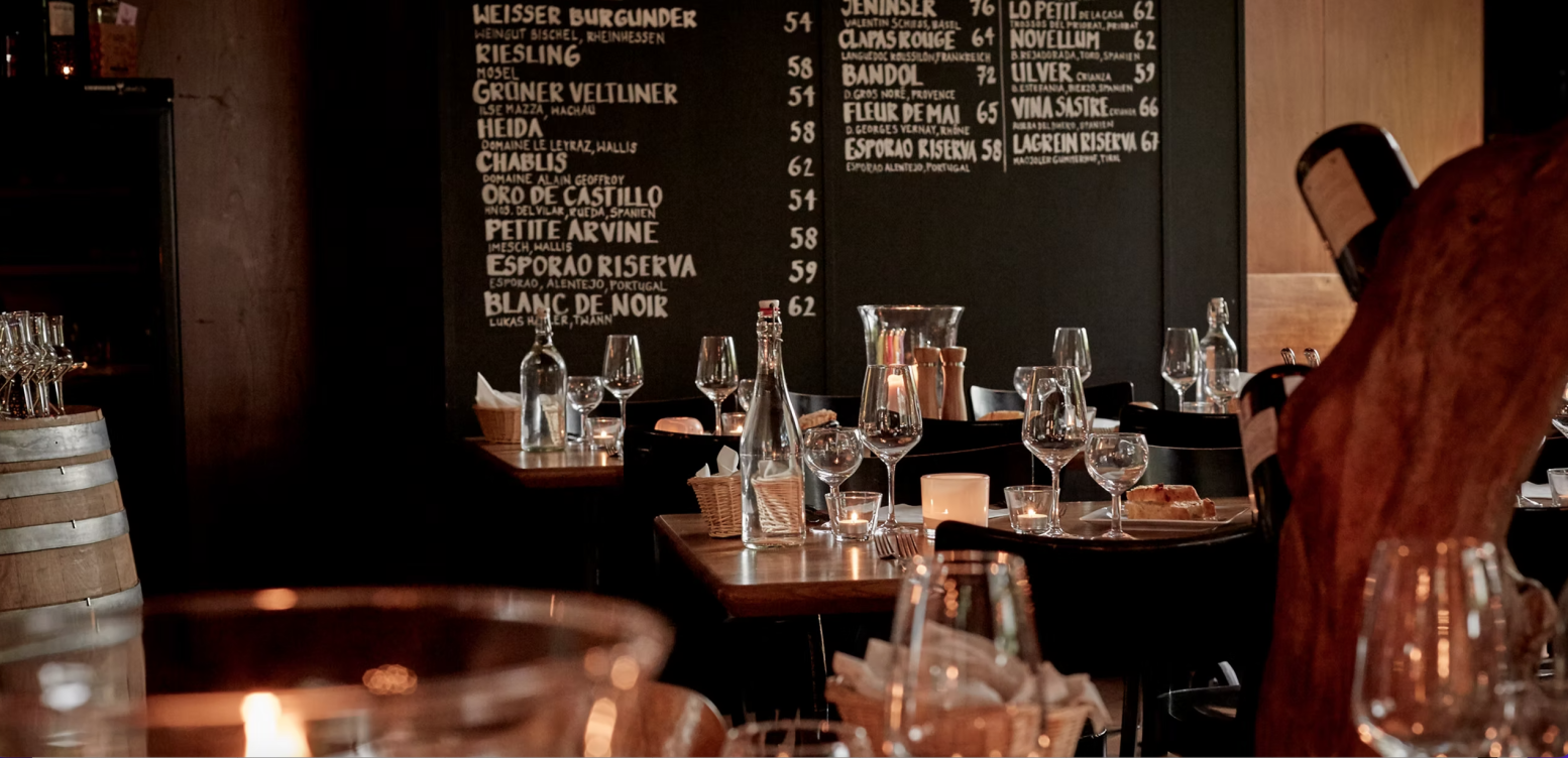 Un restaurant confortable avec un éclairage tamisé, des tables vides avec des verres à vin et des bouteilles, des bougies et une grande table avec la carte des vins en arrière-plan.