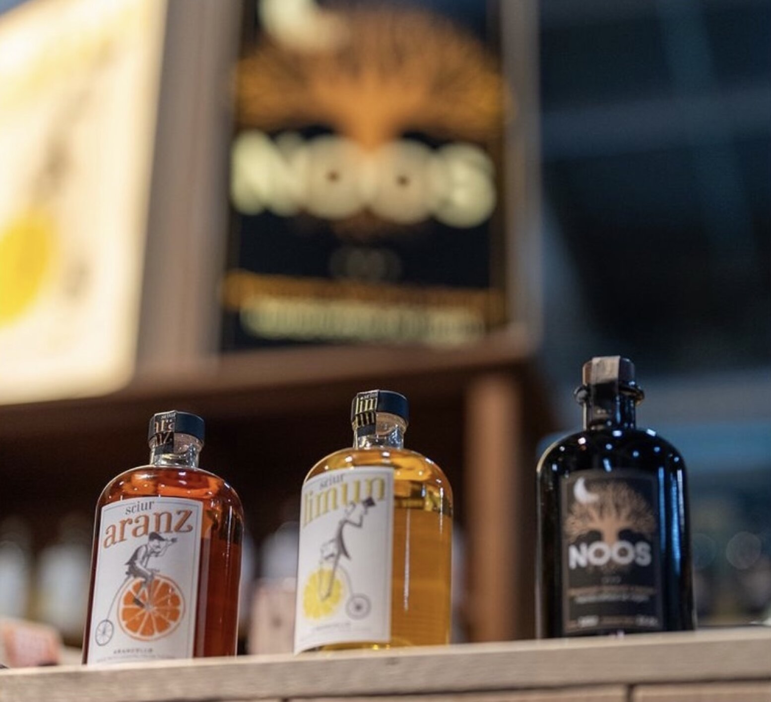 Trois bouteilles de spiritueux aromatisés sont posées sur une étagère en bois, étiquetées Aranz, Limun et NOOS. En arrière-plan, on aperçoit un panneau flou avec le logo NOOS et un arbre abstrait.