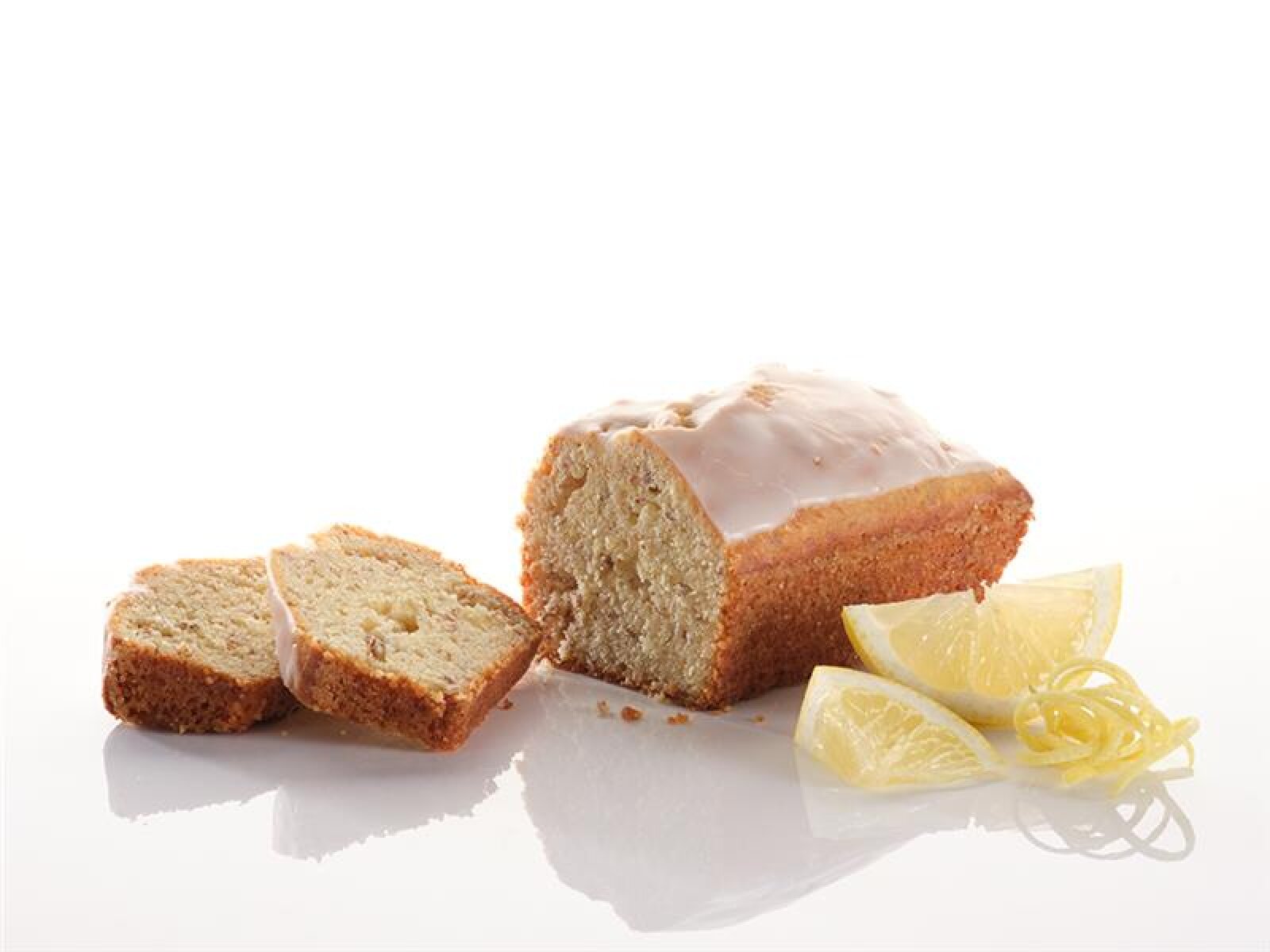 Ein glasierter Zitronenkuchen, von dem zwei Scheiben abgeschnitten und mit Zitronenspalten und Zitronenschale auf eine weiße Fläche gelegt werden.