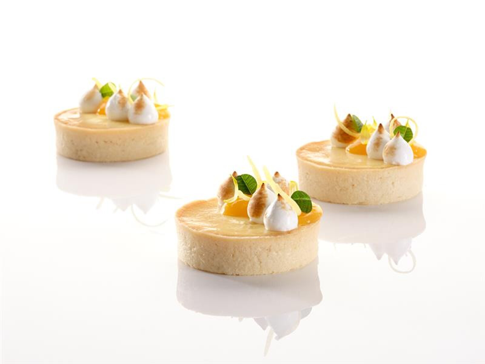 Drei Zitronentörtchen mit goldener Kruste werden mit Sahnetupfern, gerösteten Baiserspitzen und grünen Mikroblättern auf einer glänzend weißen Oberfläche angerichtet.