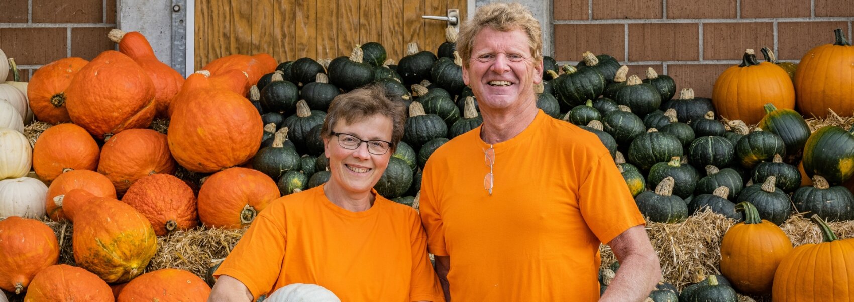 Deux personnes souriantes, vêtues de chemises orange assorties, se tiennent devant un grand étalage de citrouilles et de potirons de différentes couleurs et tailles, disposés sur des bottes de foin devant un mur de briques.