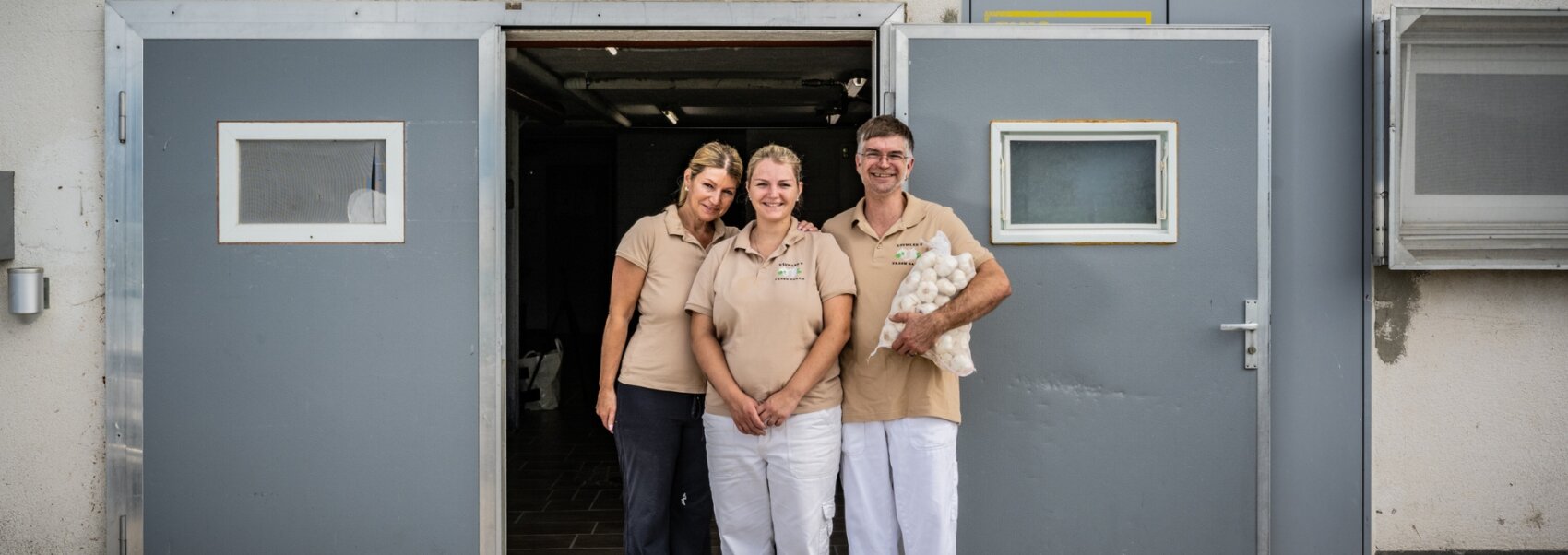 Trois personnes en chemise beige et pantalon blanc se tiennent debout, souriantes, devant de grandes portes doubles grises ; une personne tient un paquet d'objets blancs, peut-être des serviettes ou des œufs. L'environnement semble être un site industriel ou un lieu de travail.
