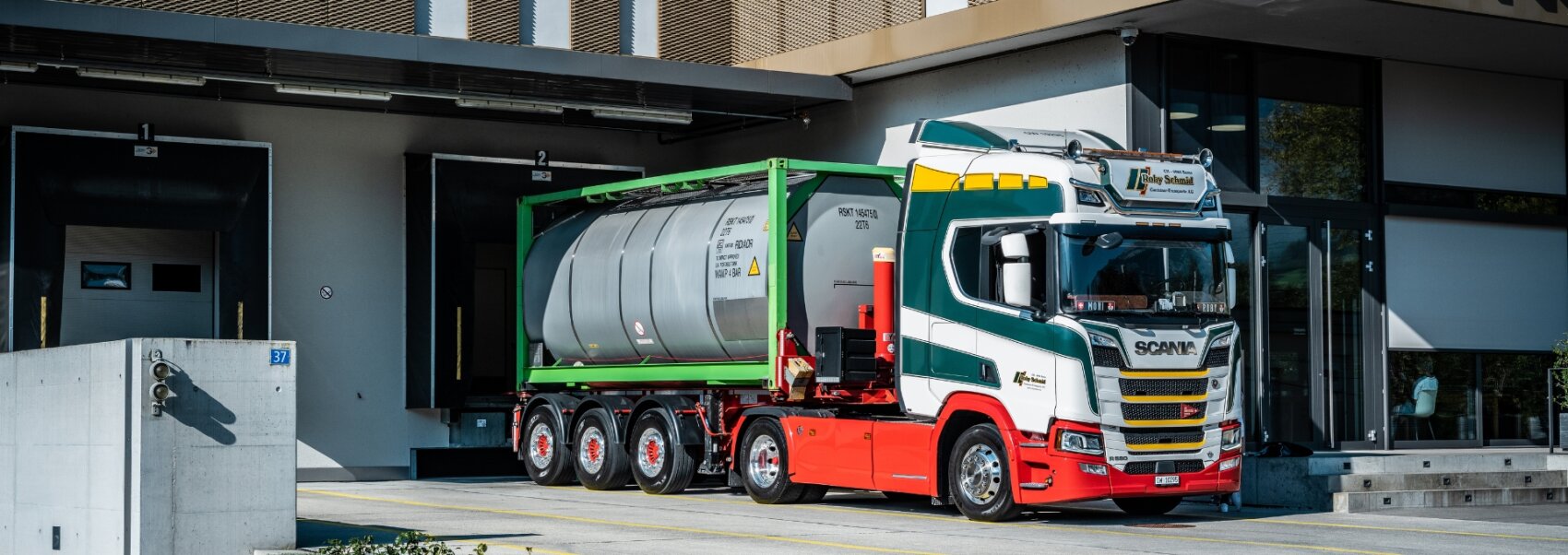 Un grand camion Scania blanc et rouge avec un cadre vert est garé sur un quai de chargement et transporte un conteneur-citerne cylindrique devant un bâtiment industriel moderne.